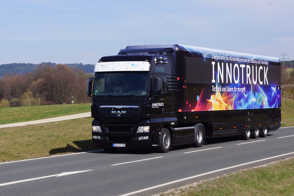 Ein schwarzer Truck mit bunter Beklebung und der weißen Aufschrift 'InnoTruck' fährt über eine Landstraße.