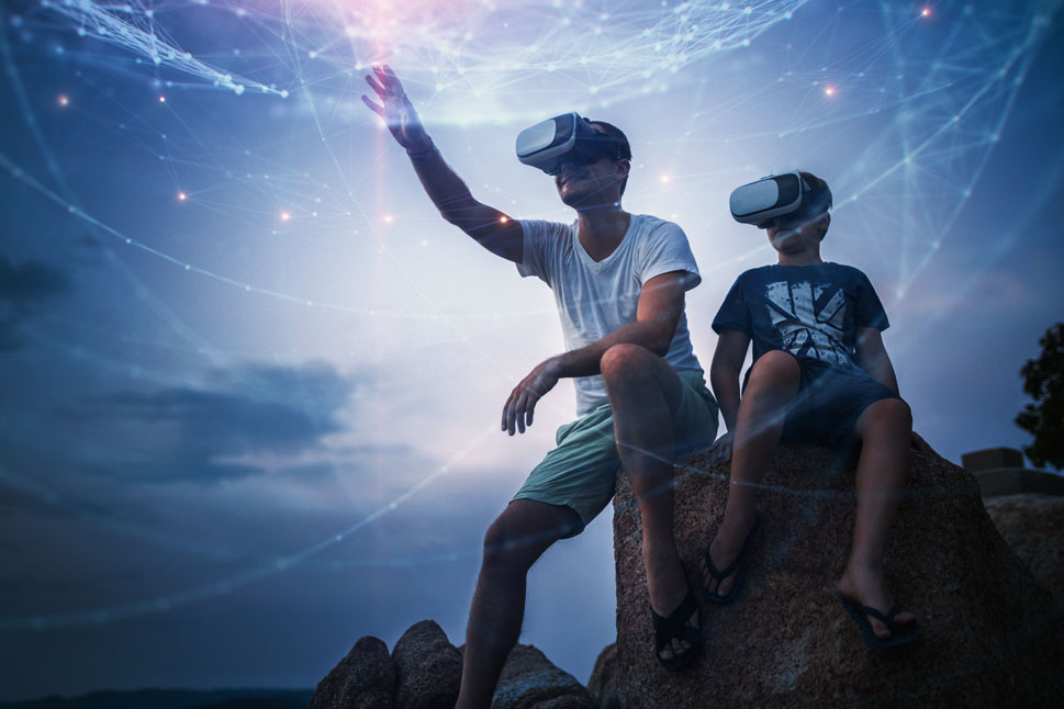 Vater und Sohn sitzen mit Virtual Reality-Brillen vor einem Nachthimmel auf einem Felsen.