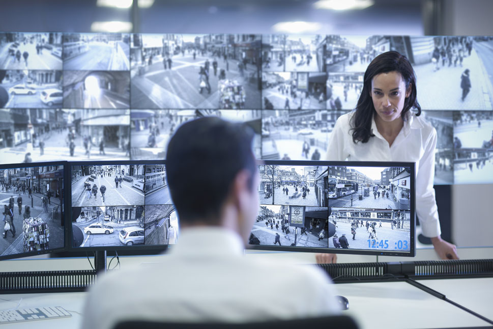 Ein Mann sitzt mit dem Gesicht zu mehreren Monitoren mit Überwachungsbildern. Eine Frau steht ihm gegenüber.