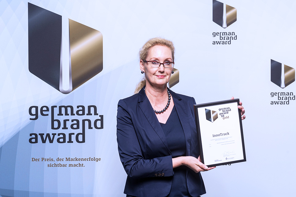 Eine blonde Frau hält eine Urkunde in ihrer Hand und lächelt damit in die Kamera. Sie steht vor einer Wand mit den Logos des German Brand Awards.