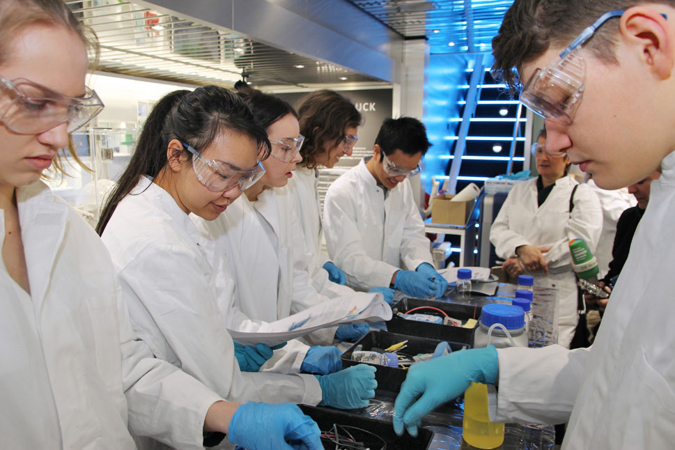 Schülerinnen und Schüler in Laborkleidung stehen gegenüber an einem Tisch und sortieren Laborutensilien.