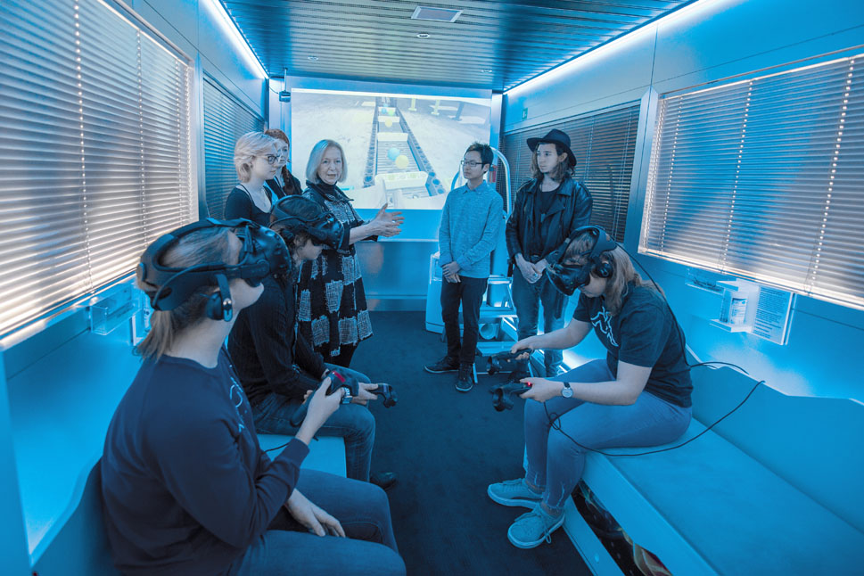 Drei Jugendliche sitzen auf gegenüberliegenden Bänken und spielen ein Virtual-Reality-Spiel. Die Ministerin steht mit weiteren Jugendlichen daneben und beobachtet die Szene.