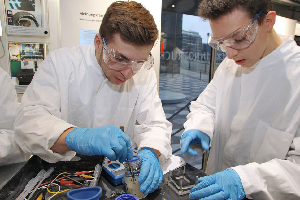 Zwei Schüler in Laborkleidung stehen an einem Tisch und bauen aus Kabeln, einem Gestell und Platten eine Redox-Flow-Batterie zusammen.