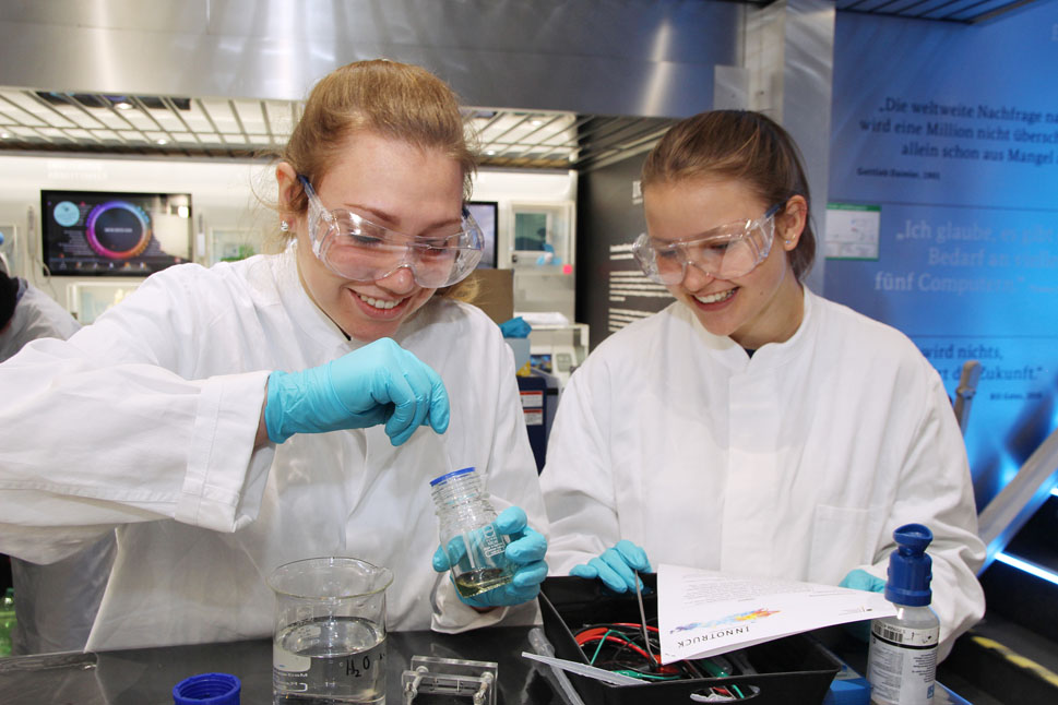 Zwei Schülerinnen in Laborkleidung stehen an einem Tisch und halten Reagenzgläser in der Hand.