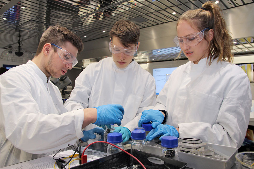 Zwei Schüler und eine Schülerin in Laborkleidung arbeiten zusammen mit verschiedenen Labormaterialien.