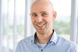 Bild zeigt einen lächelnden Mann mit kurzen Haaren und hellblauem Hemd vor einem hellen Hintergrund