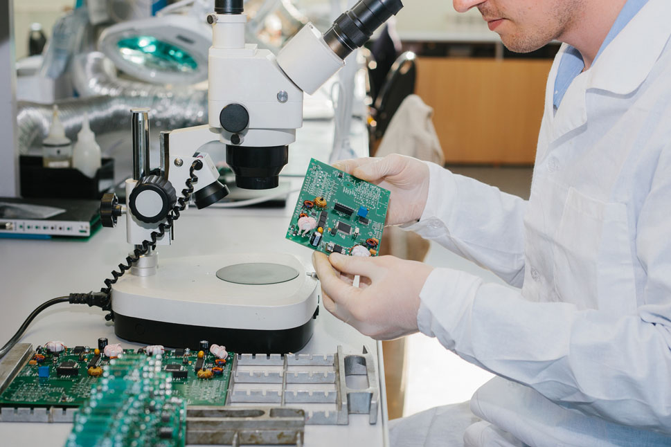 Bild zeigt einen Mann im weißen Laborkittel, der vor einem Mikroskop sitzt und auf eine Computerplatine in seiner Hand blickt