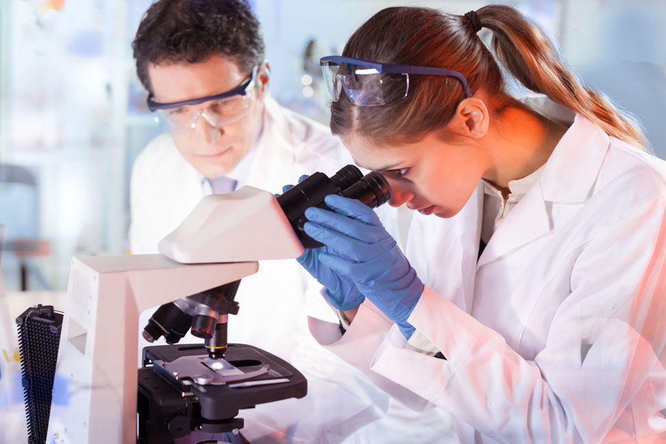Bild eine Wissenschaftlerin im weißen Laborkittel, die durch ein Mikroskop blickt. Ein Kollege beobachtet das Geschehen.