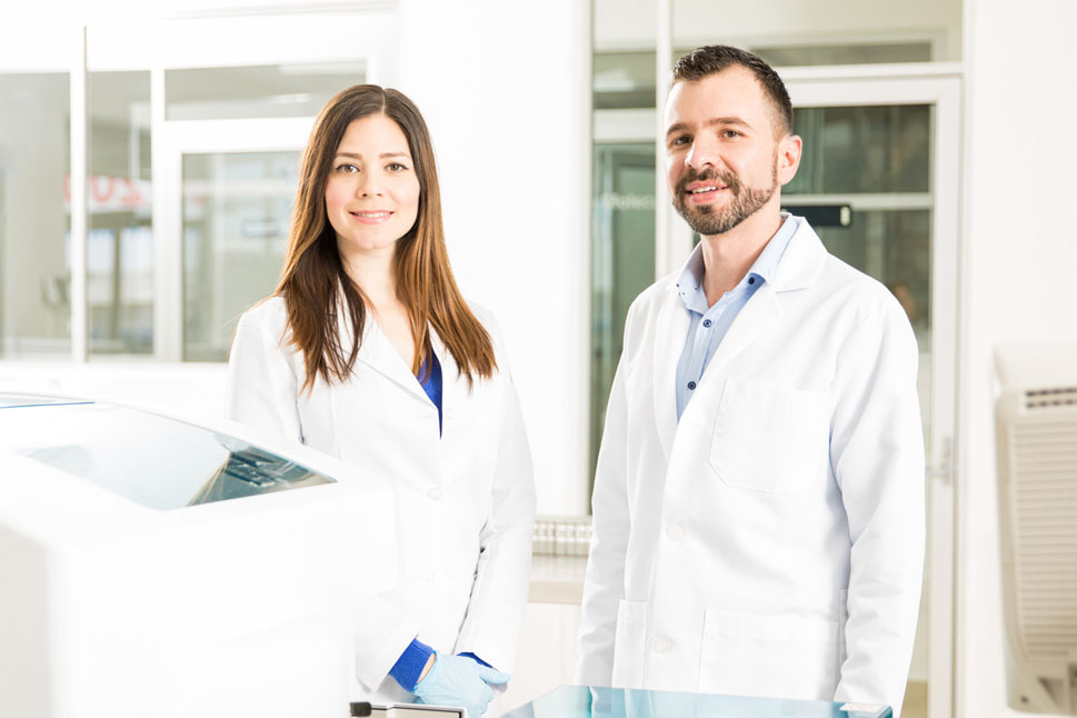 Bild zeigt eine Frau und einen Mann in weißen Laborkitteln in einer hellen Laborumgebung stehen, die in Richtung des Betrachters blicken