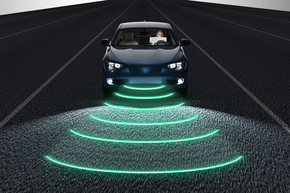Bild zeigt ein Fahrzeug auf einer leeren Straße. Anhand einer wellenförmigen Visualisierung an der Fahrzeugfront und einem lesenden Fahrer, erkennt man, dass es ein autonomes Fahrzeug ist.