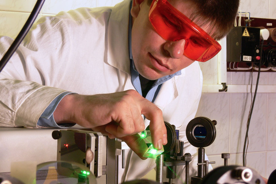 Wissenschaftler in weißem Kittel und mit roter Schutzbrille beugt sich über einen Versuchsaufbau mit einem grünen Laserstrahl