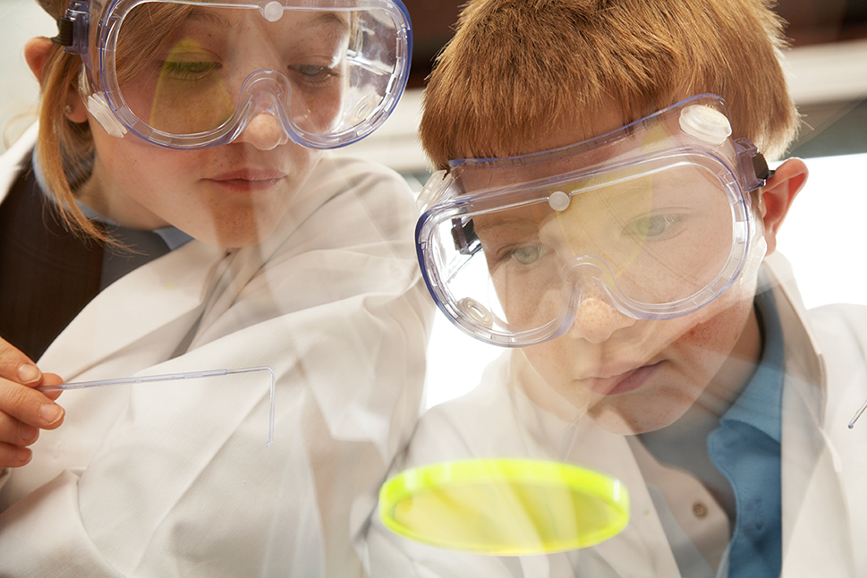 zwei Schüler mit Schutzbrillen und weißen Laborkitteln experimentieren mit einer leuchtenden Substanz in einer Petrischale