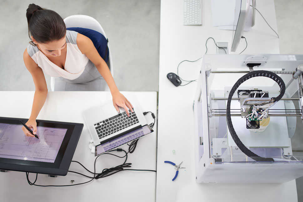 Bild zeigt Frau, die an einem Schreibtisch mit Notebook, Touchscreen und 3D-Drucker arbeitet